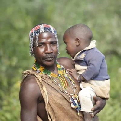 A Tanzanian father wearing traditional ornaments carries his son.
