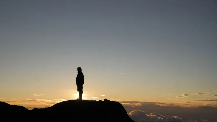Silhouetted figure stands at the top of a peak in Tanzania with sun illuminating the clouds that hug the mountains.