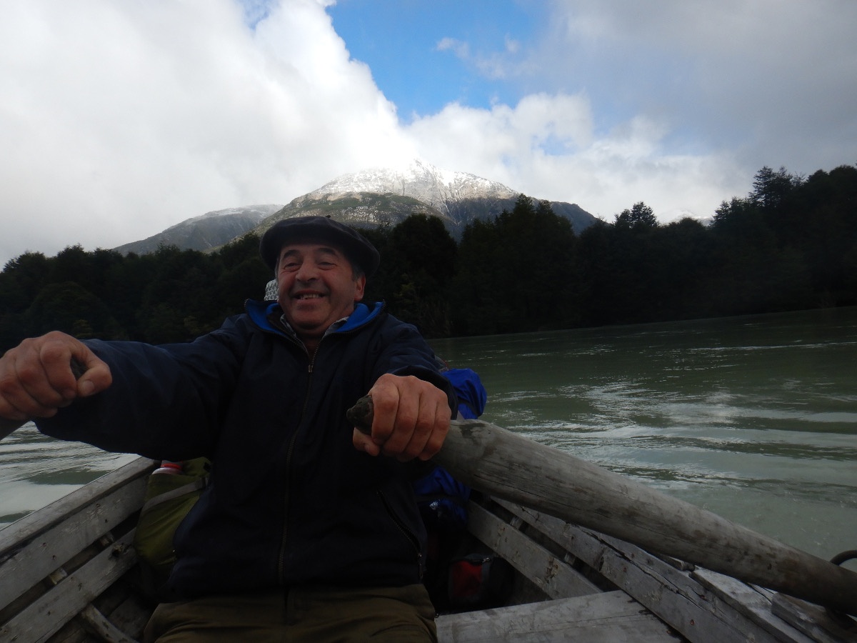 person rows a wooden boat on a lake with dark trees and snow-capped mountain in the background