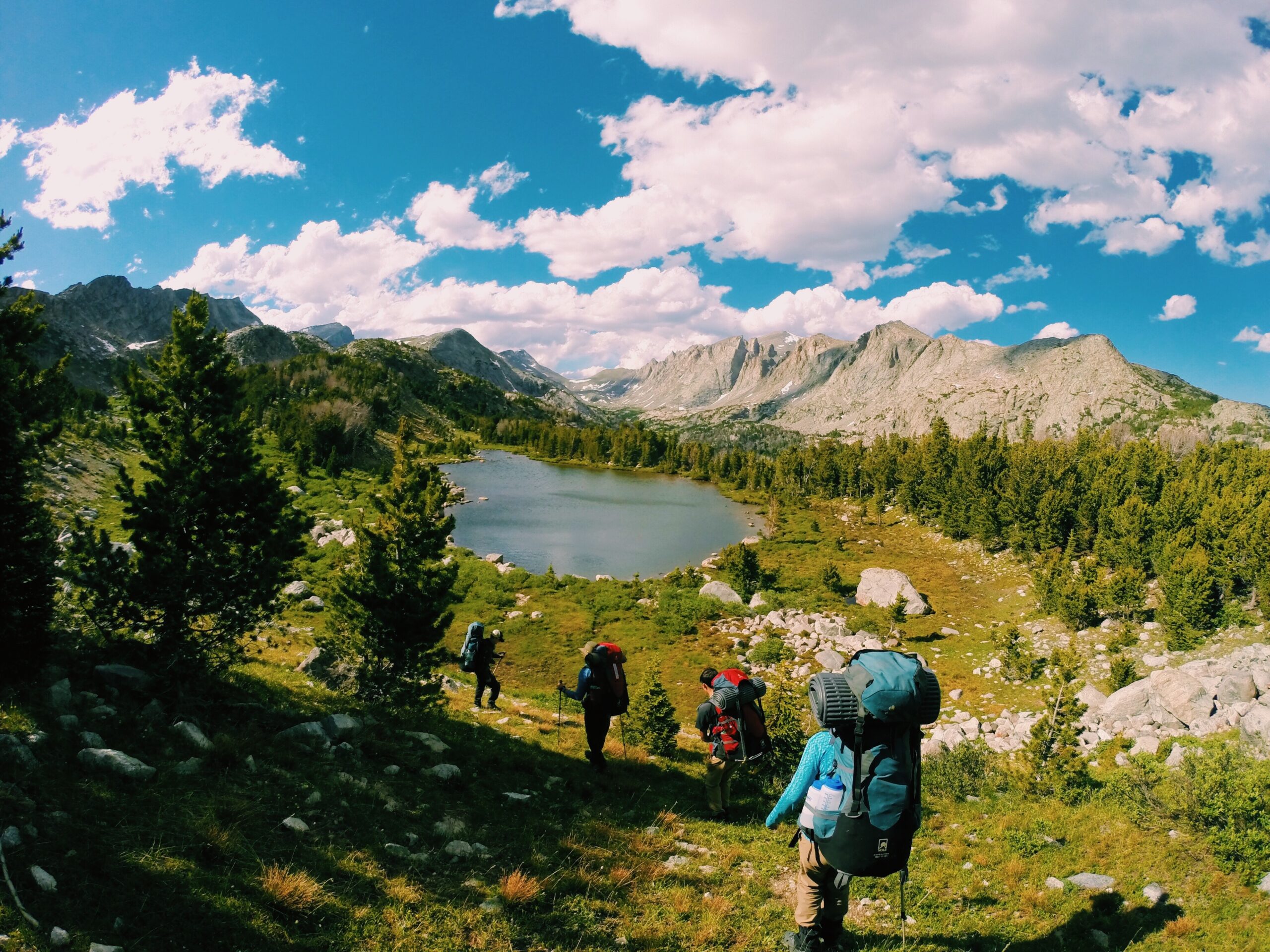 students with backpacks hike down a green slope toward an alpine lake in the Wind River Range