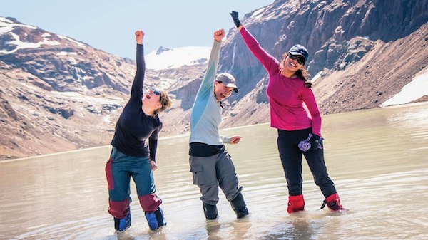 Three smiling NOLS participants stand in shallow mountain lake and raise arms in celebration