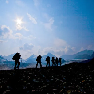 Silhouetted students backpacking along scree with snowy mountains and blinding sun in the distance. Credit: Madhu Chikkaraju