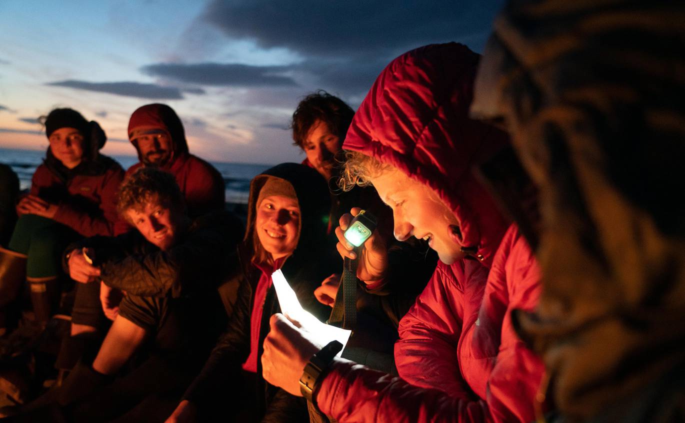 A student uses a headlamp to read off a piece of paper during a group meeting around the campfire.