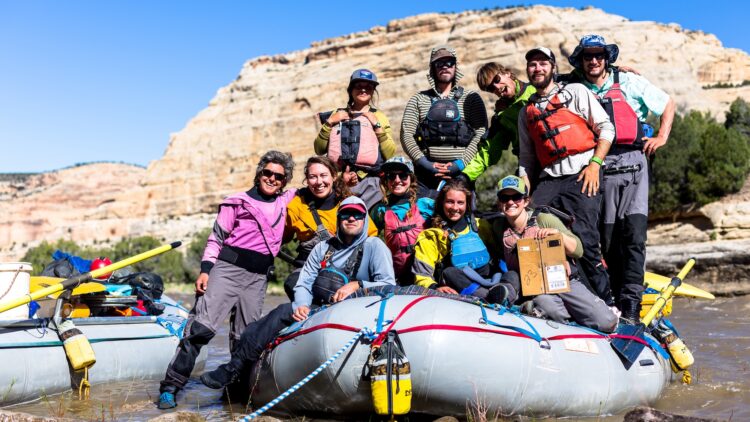A group of ten people pose on an inflatable raft. They are wearing PFDs and drysuits and all are smiling.