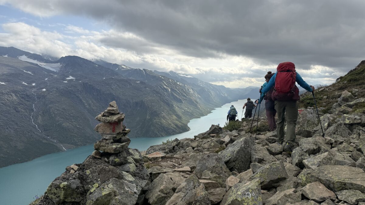 Group hikes on rocky terrain with long, narrow lake to their left and mid-sized cairn also to left.