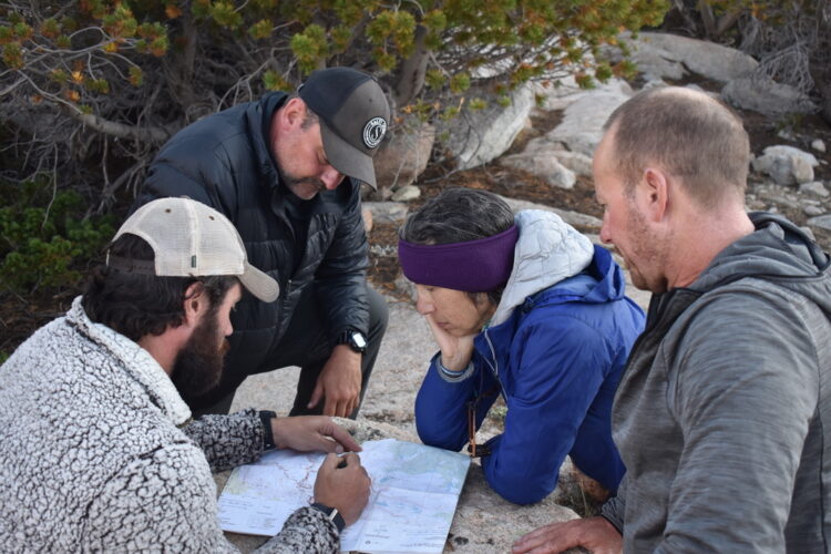 A group of four people sit on a large granite ledge looking at a map. A man in the left foreground using a pen to show something on the map.