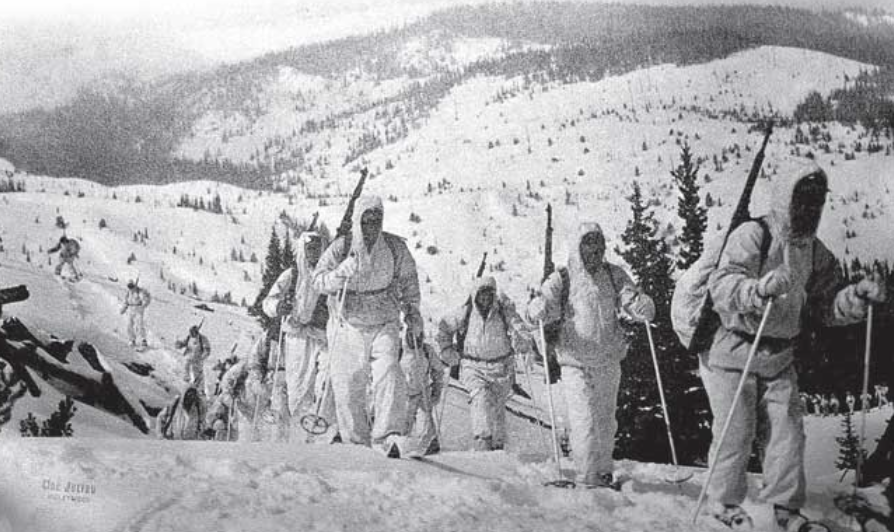 Members of 10th Mountain Division, dressed in white camouflage and carrying weapons, move through the mountains.