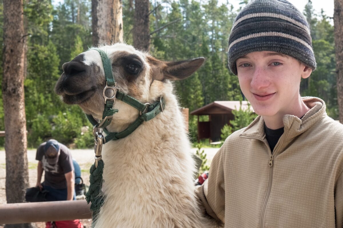 Boy wearing a knit cap, posing with a brown and white llama, as they prepare to head into the mountains.