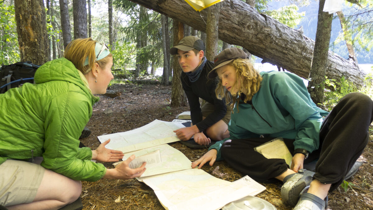 Two students on right examine notes and maps with their course instructor, on left, who is wearing a bright green jacket They are in a wooded area with water in the background