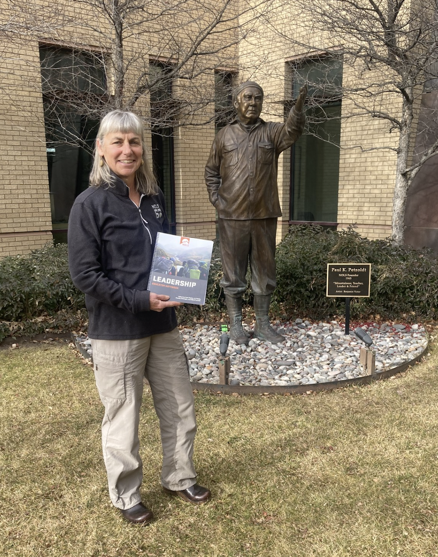 Photo of Shannon Rochelle, NOLS Expedition Education Director, holding a copy of the new (published in late 2025) Leadership Educator Notebook. She stands in front of the statue of Paul Petzoldt, which is on the small lawn in front of HQ.