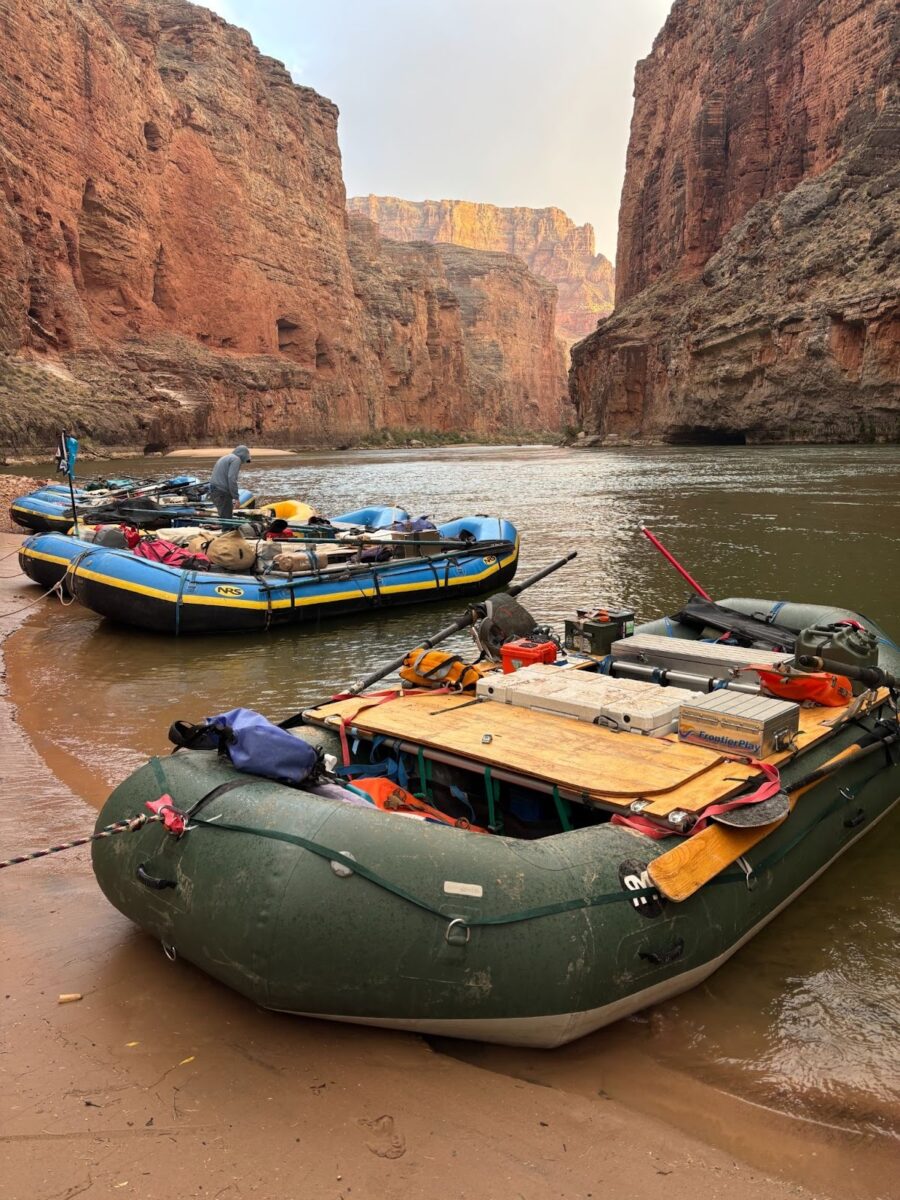 Three rafts are docked on a beach on the Colorado River in the Grand Canyon. A man on the center raft appears to be looking for something or checking something. The river is surrounded by steep red cliffs.