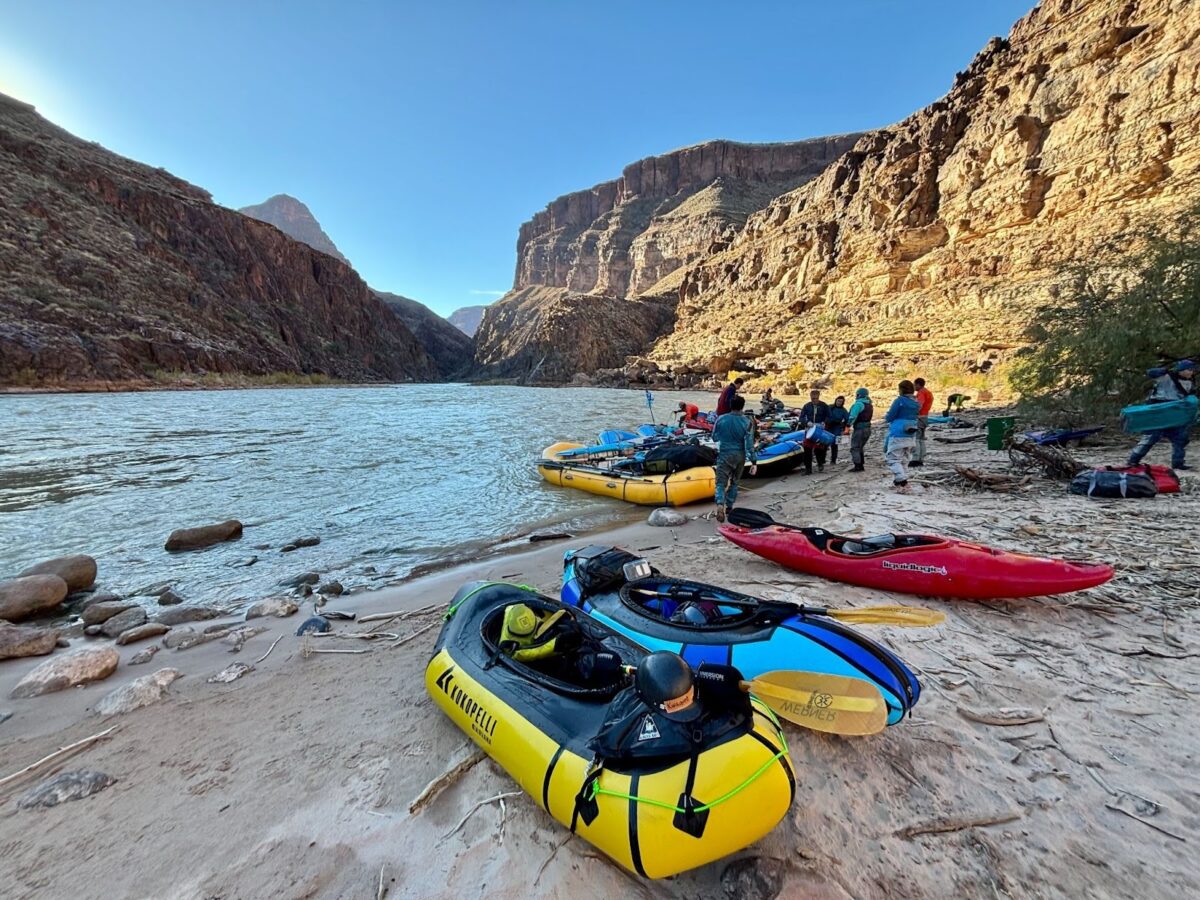 A group is standing by their rafts and a kayak, several of them beached on the banks of the Colorado River in the Grand Canyon. Sunlight is striking the cliffs on the right side, while blue skies are visable above.