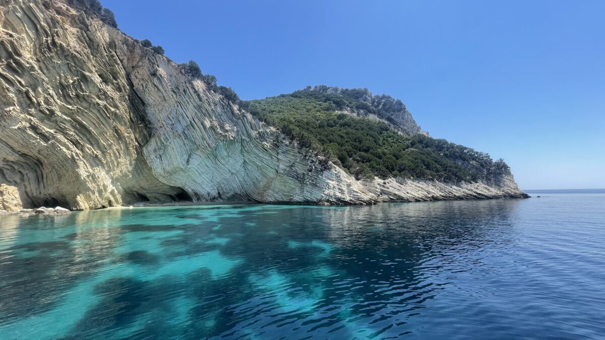 Shades from aqua to deep blue in the water with a cliff, partially covered in green trees to the left.