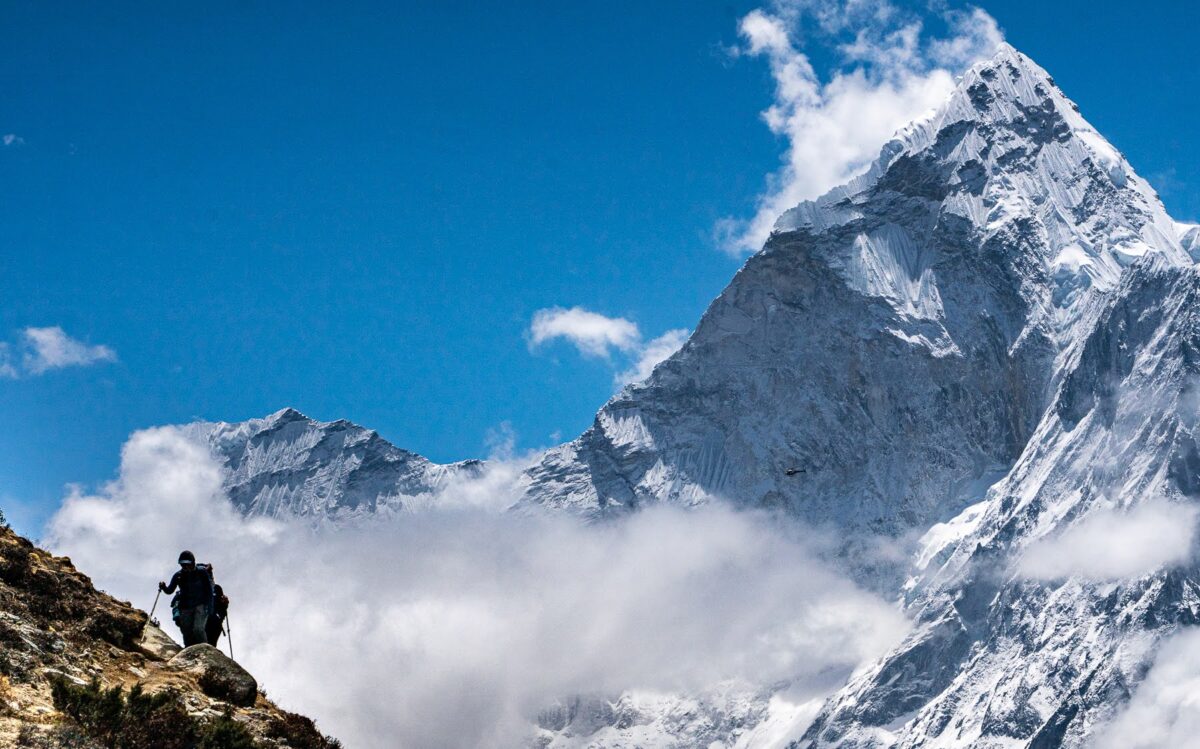 Team members are visible on a path in the right side of the photo. Everest is in the background, surrounded by blue skies and filmy white clouds.