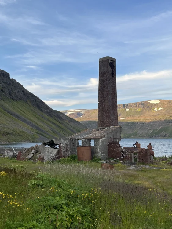 an abandoned brick building with a tall chimney. behind it is a lake and mountains. 