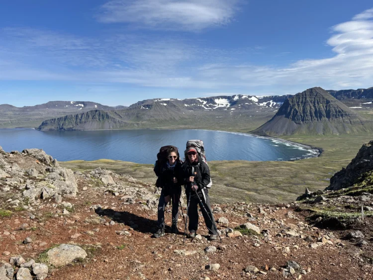 Two people with backpacks on pose in front of a large body of water. The sky behind them it blue. 
