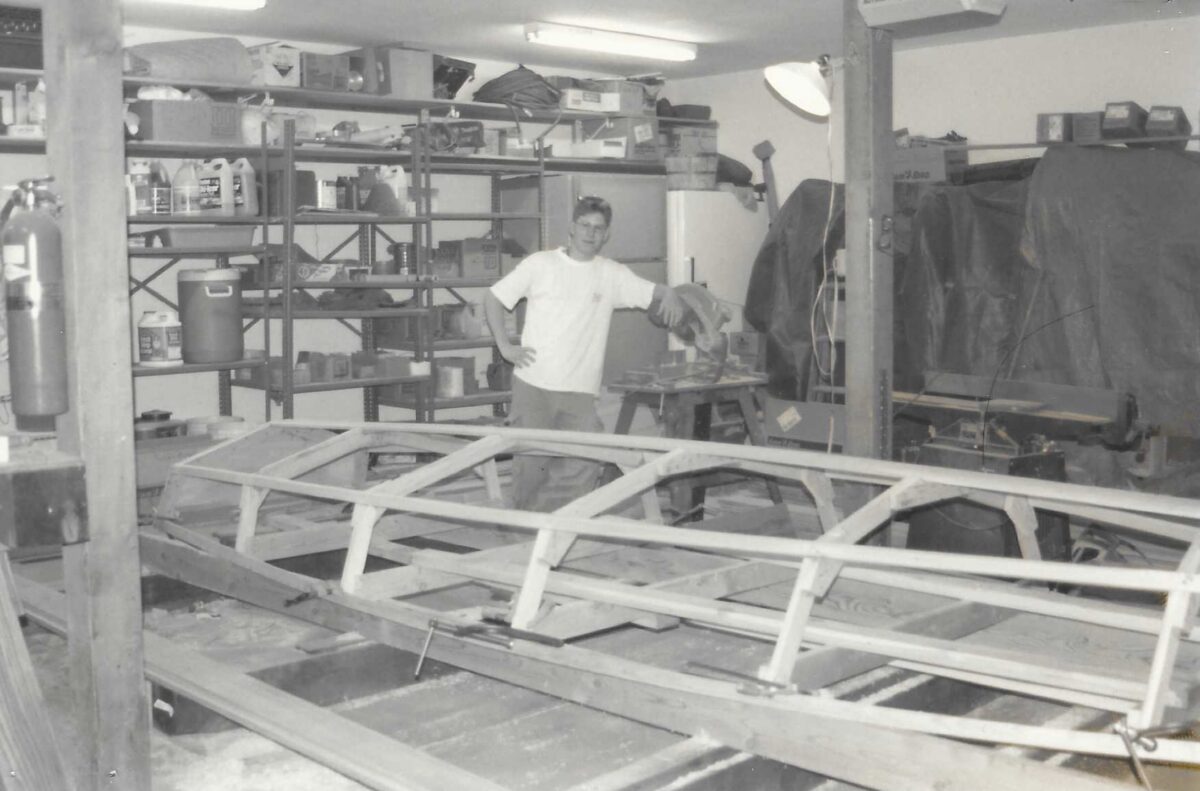 B/W photo of the skeleton of Nick's sailboat in the garage as he and his dad built it. Nick stands behind it.