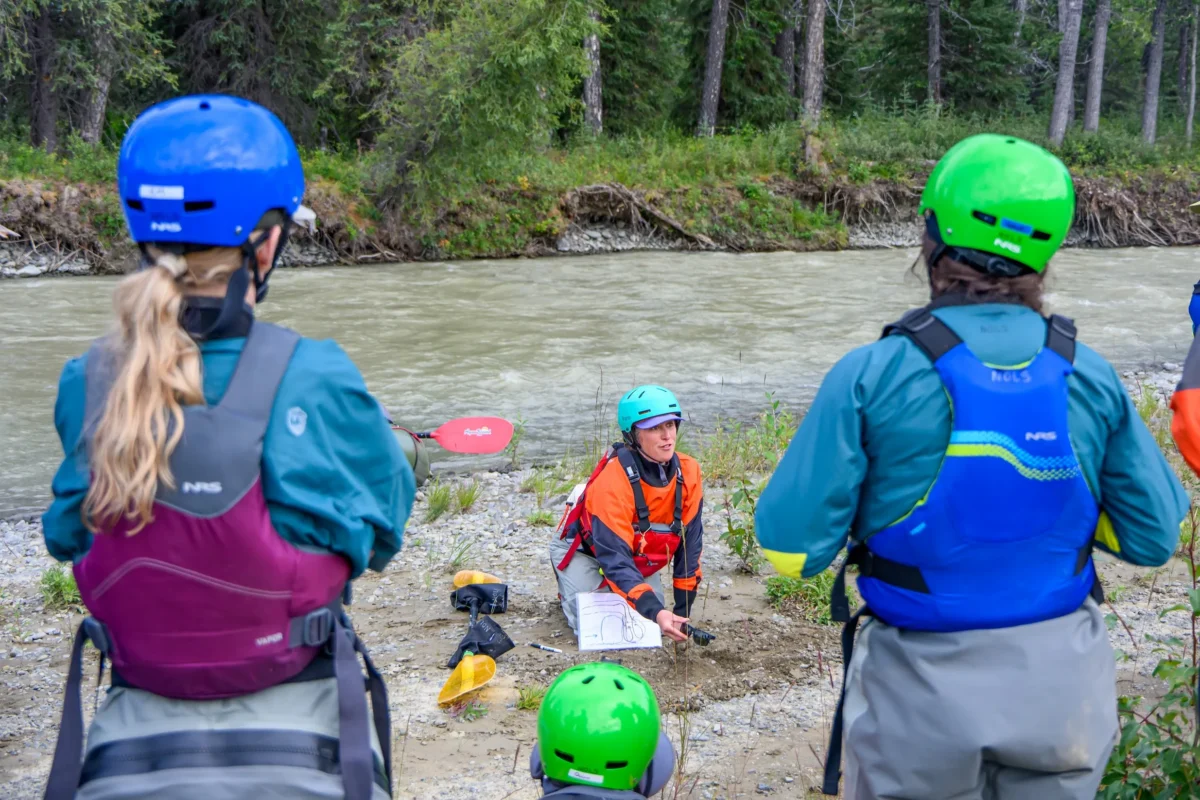 NOLS whitewater kayaking instructor in an orange dry suit and teal helmet teaches a group of students on a rocky riverbank in Alaska, with participants wearing colorful safety helmets and PFDs while reviewing a map beside a glacial river bordered by dense evergreen forest.