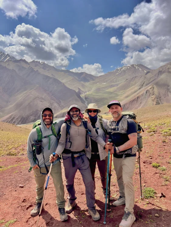 Four hikers pose with their backpacks and trekking poles with towering mountains in the background.