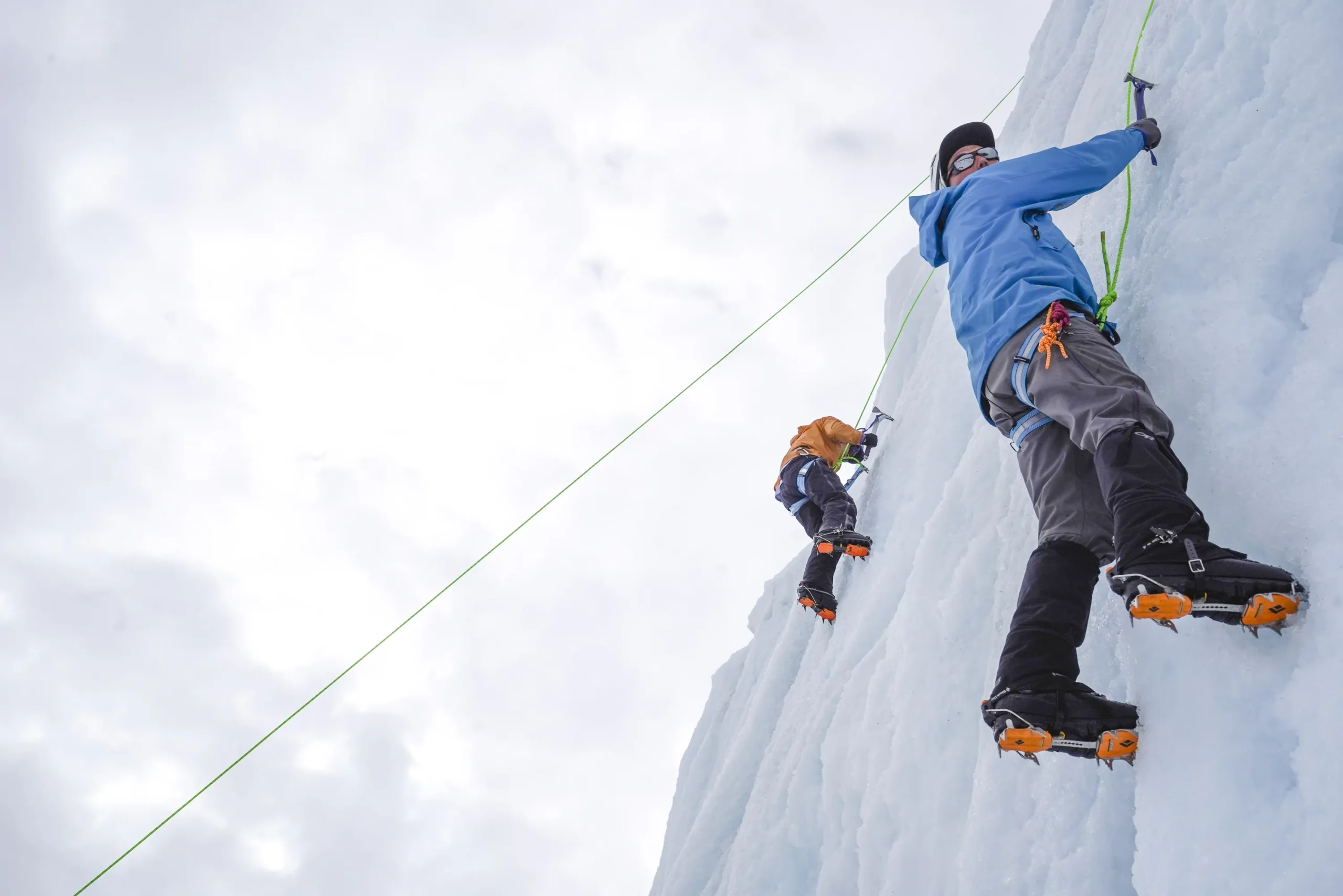 A person in a blue jacket climbs a wall of ice. They have crampons on their boots and are holding ice axes in their hands.