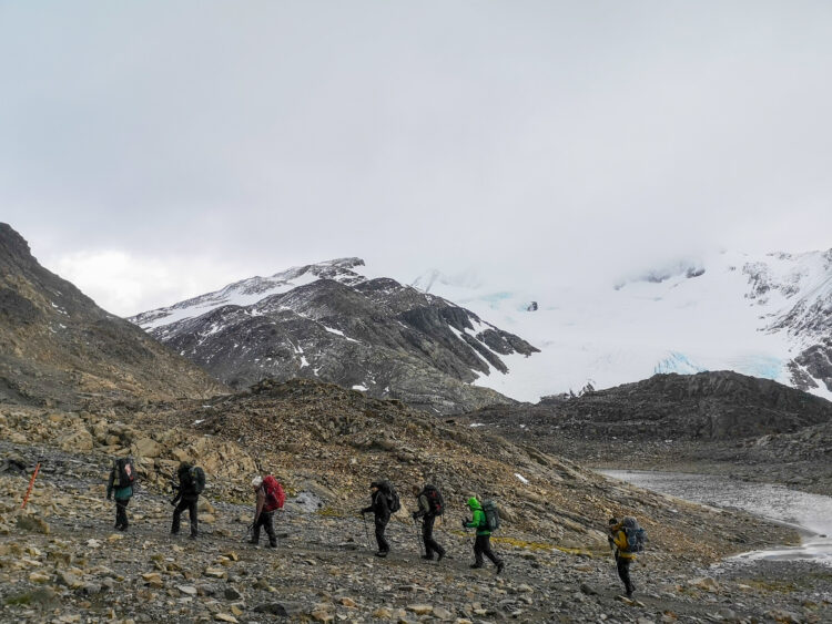 7 people walk across a rocky mellow mountain slope. It's a grey day and there are glaciers in the background.
