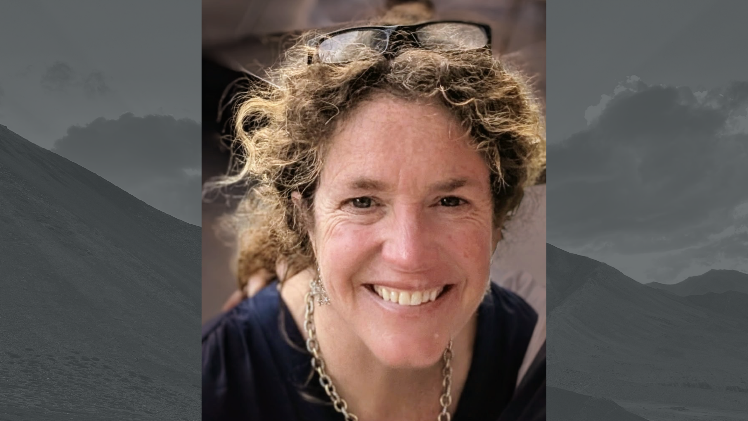 a woman with curly hair in a ponytail and glasses on her head smiles for the camera.