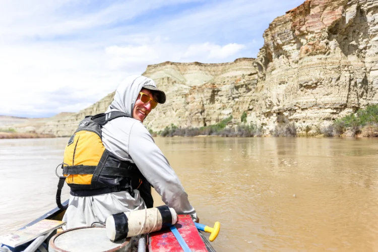 a person wearing a grey hoodie, a hat, sunglasses, and a yellow PFD sits on a boar in a river. they are smiling at the photographer.