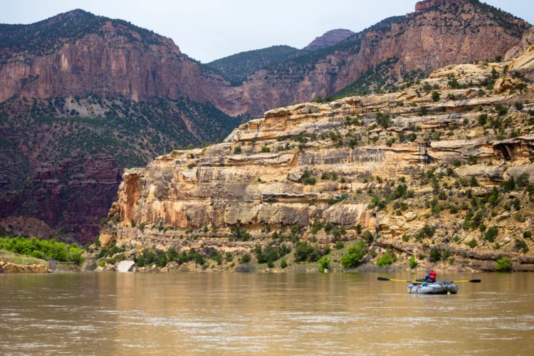 a blue inflatable raft drifts on a brown river between red canyon walls. 