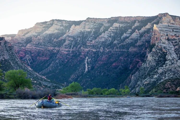 a person rows an inflatable raft on a river. There is a tall canyon wall in the background.