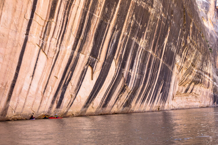 a large wall of red and black rock rises above a river. three kayaks float beside the base of the wall