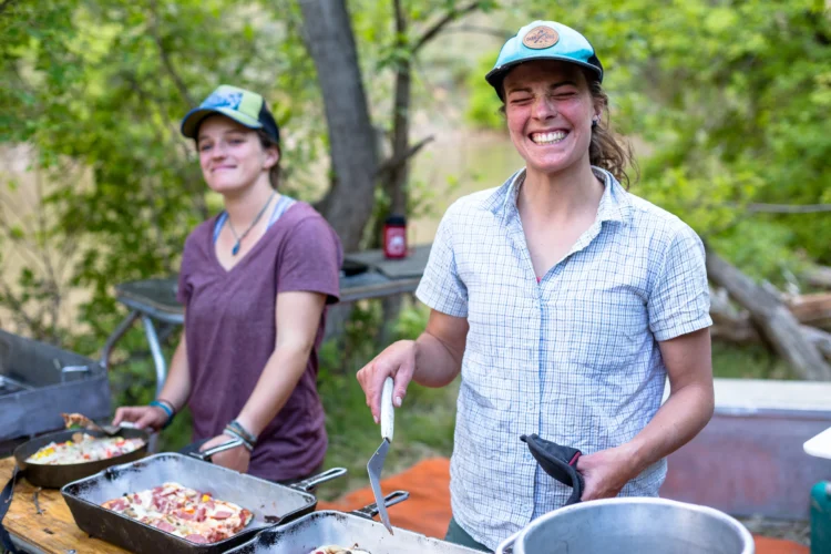 Two women stand at a table outdoors. They are both wearing trucker hats and smiling at the camera. One is serving food to people. 