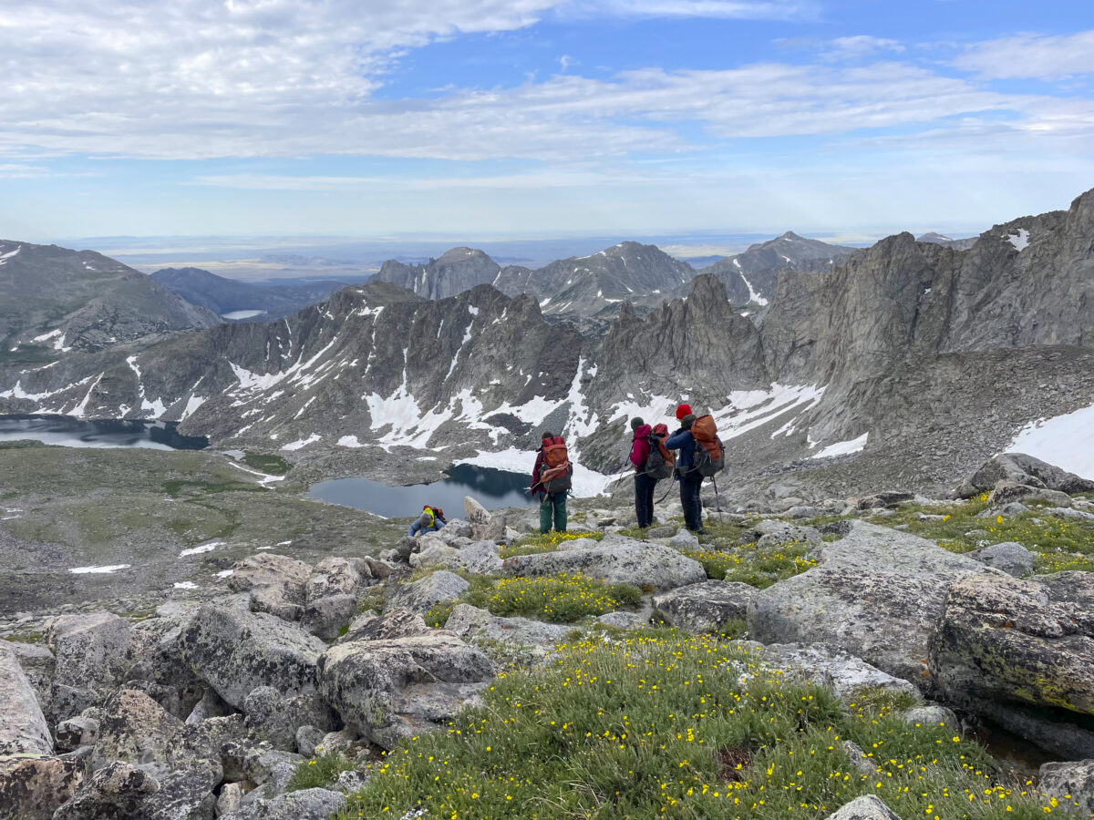 three people wearing backpacks stand on a mountain pass looking out at granite peaks.