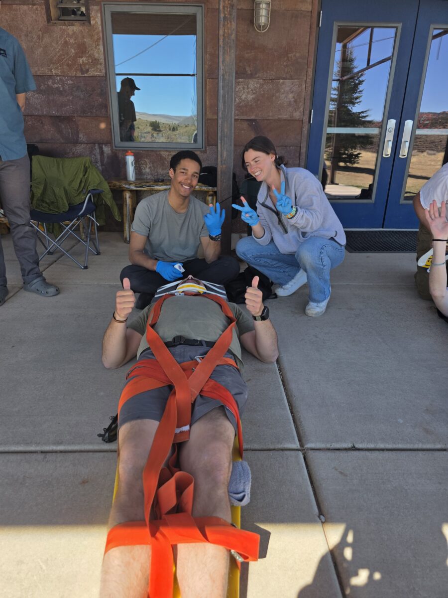 two people wearing blue medical gloves pose for the camera. theres is a man on a backboard giving thumbs up to the camera.