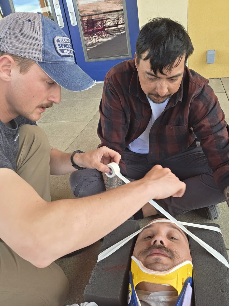 a man's head is cradled by blocks of foam that are being taped in place. he is also wearing a neck collar. two men are caring for him.