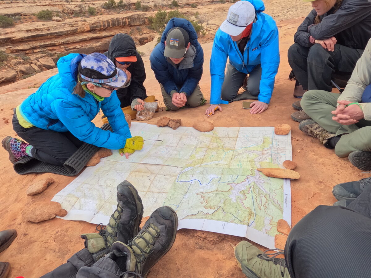 a group of eight people in a circle look at a map that is resting on red slickrock. the people are all wearing outdoor clothes and ball caps.