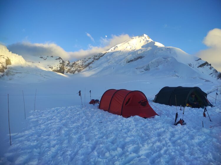 two mountaineering tents are set up on a snow field between two mountain peaks
