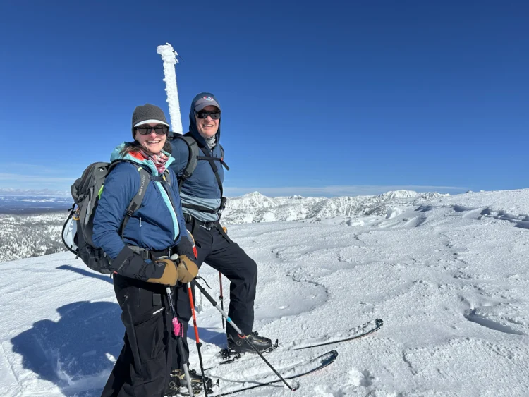 two people wearing ski smile at the camera on a snowy ridge. the sky is blue behind them