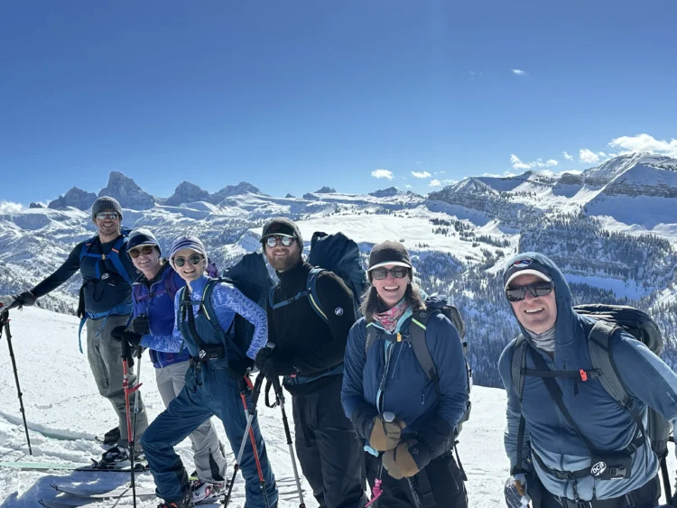 six people wearing skis and winter apparel, including sunglasses, smile at the camera. there are lots of mountains in the background and the sky is blue