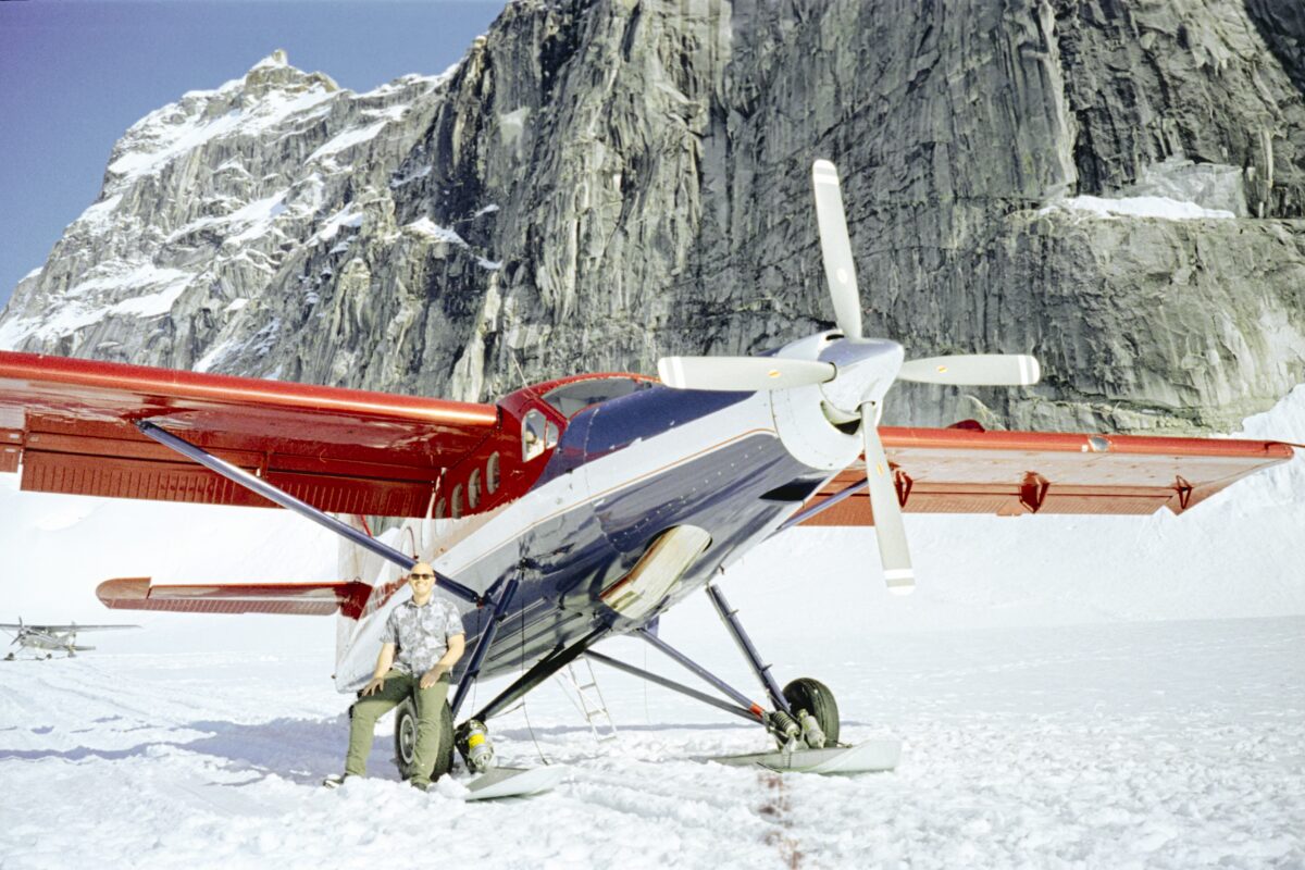 Asa leans against his plane on a snow-covered patch of land while mountains tower behind him.