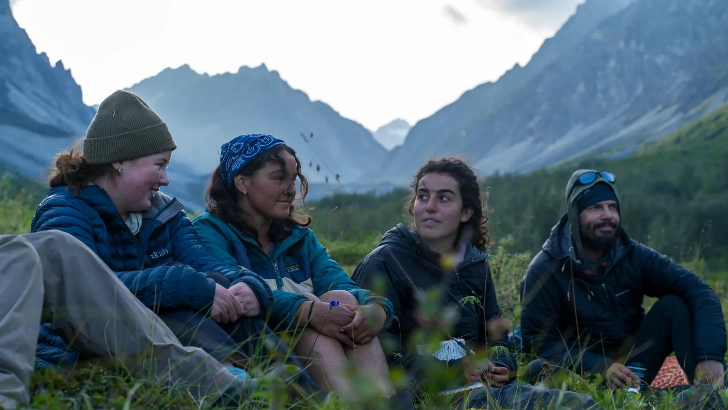 four people sitting outside in front of large mountains smile at one another