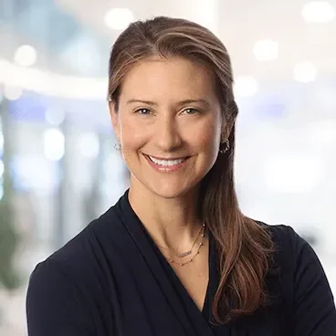 A woman with medium-length brown hair poses against a blurry background. She is smiling at the camera.