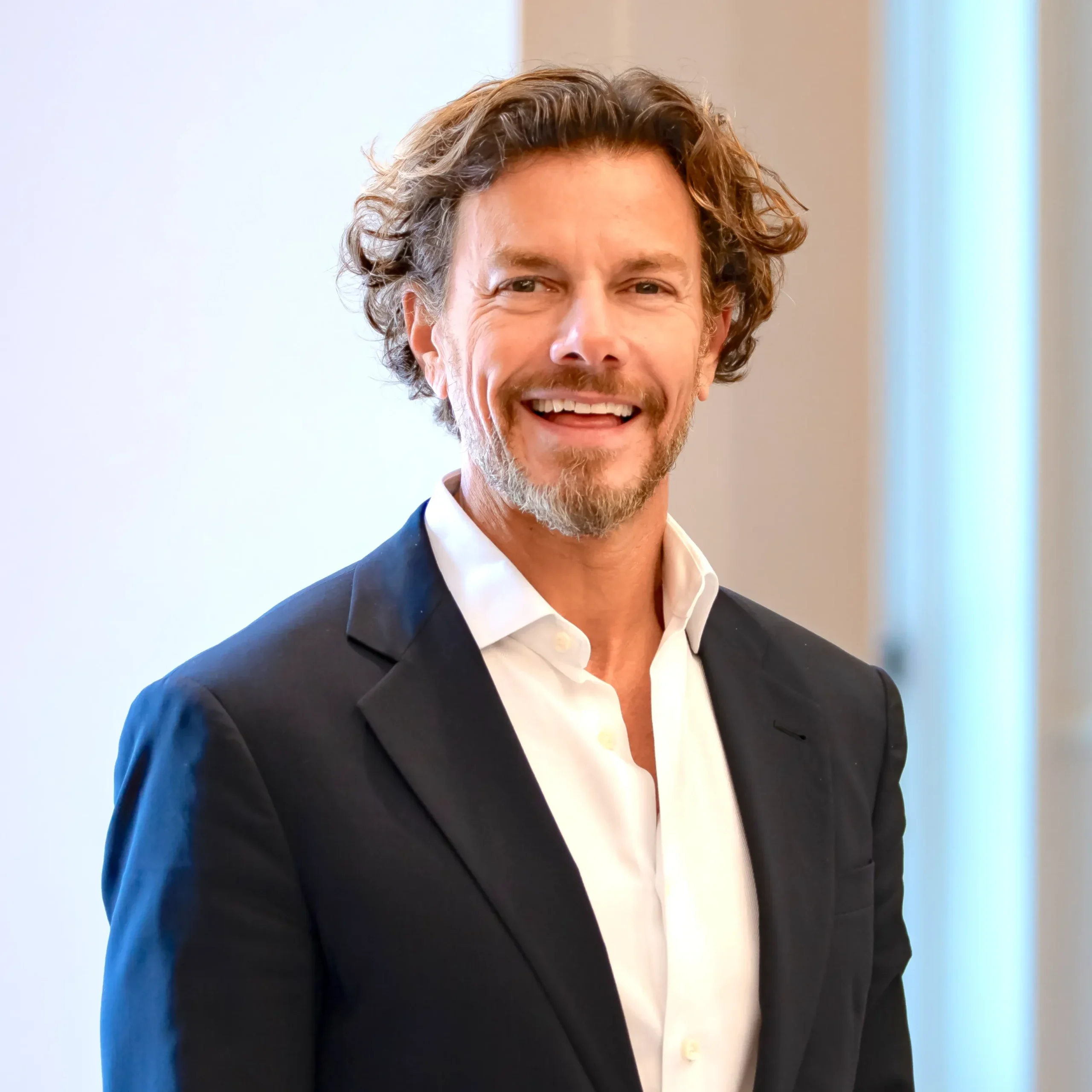 A man with wavy brown hair and a blue suit jacket smiles at the camera.