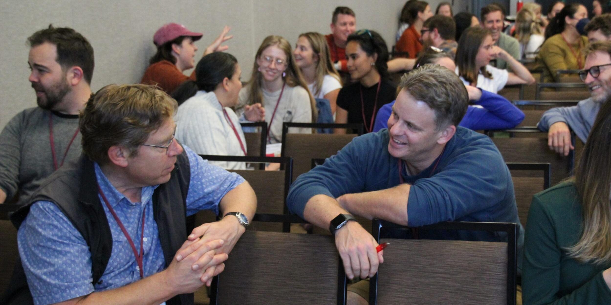 two men in a classroom talk to one another across a chair. many people in the background are engaged in conversation